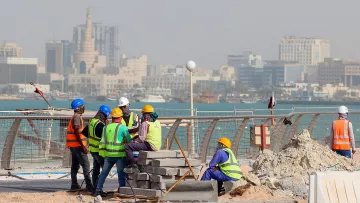 L'image montre un groupe de travailleurs sur un chantier de construction près de la mer. Ils portent des vêtements de sécurité, comme des gilets réfléchissants et des casques. En arrière-plan, on peut voir une ville avec des bâtiments modernes et des structures élevées. L'ambiance semble occupée, et le chantier est environné de matériaux de construction, avec des blocs empilés. La mer est visible au loin, ajoutant un cadre pittoresque à la scène.