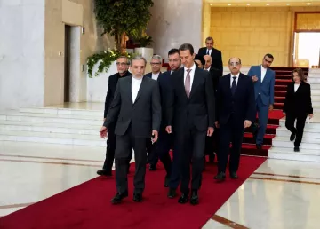 L'image montre un groupe de personnes marchant sur un tapis rouge dans un hall d'entrée élégant. À l'avant, deux hommes semblent en conversation, tandis que d'autres personnes les suivent, allant d'une manière formelle. L'environnement est spacieux avec des murs clairs et une décoration soignée, indiquant un lieu officiel ou diplomatique.
