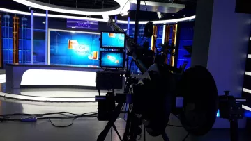 L'image montre un studio de télévision moderne. On peut y voir des lumières bleues qui éclairent l'espace, avec un fond décoratif et des écrans de présentation. À l'avant-plan, il y a une caméra sur un trépied, ainsi que des équipements techniques, ce qui indique que le studio est préparé pour une émission en direct ou un enregistrement. L'ambiance semble professionnelle et bien équipée pour la diffusion.