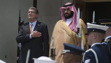 در این تصویر، دو مرد ایستاده‌اند. مرد سمت چپ با یک کت و شلوار تیره و پیراهن روشن دیده می‌شود و دستانش را بر روی سینه‌اش گذاشته است، که نشان‌دهنده احترام یا تقدیر است. مرد سمت راست در لباس رسمی عربی و با عمامه‌ای که به طور خاص مرتب شده، stands. در پس‌زمینه، گروهی از سربازان در یونیفورم‌های نظامی در حال ایستادن هستند. این صحنه احتمالاً به یک مراسم رسمی یا استقبال سیاسی مربوط می‌شود.