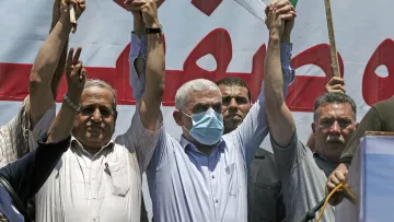 The image depicts a group of men at a public gathering or protest. They are raising their hands in the air, with some holding sticks or flags. The man at the center is wearing a mask and appears to be leading or participating actively in the event. In the background, there is a large banner in Arabic, suggesting a political or social message. The mood of the event seems to be one of solidarity or protest, with participants visibly engaged.