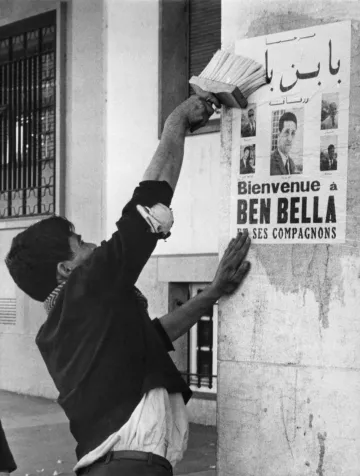 L'image montre un homme en train de coller une affiche sur un mur. L'affiche annonce un message de bienvenue à Ben Bella et ses compagnons, tout en affichant plusieurs portraits. L'homme semble concentré et utilise un outil pour bien lisser l'affiche contre la surface. L'environnement urbain suggère une période historique, probablement liée à un événement important. Les détails architecturaux et la tenue de l'homme donnent une impression de contexte social et culturel spécifique.
