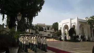 L'image montre une scène extérieure d'un bâtiment élégant, probablement un palais ou un hôtel de luxe. On peut voir une allée centrale avec un tapis rouge, entourée de verdure et de palmiers. À gauche, un groupe de personnes en uniformes formés, représentant une sorte de garde ou de cérémonie. En arrière-plan, on aperçoit un bâtiment majestueux avec des arches et des colonnes, suggérant une architecture raffinée. Le ciel est clair, ce qui ajoute à l'atmosphère solennelle de la scène.