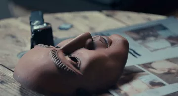 L'image montre un masque en terre cuite posé sur une surface en bois. Le masque a des traits détaillés, avec une expression neutre et des sourcils sculptés. À côté du masque, on voit une montre ou un objet similaire, ainsi qu'un magazine ouvert avec des illustrations. L'éclairage est doux, créant une ambiance contemplative autour de ces objets.