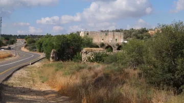 L'immagine mostra un paesaggio rurale caratterizzato da una strada curvilinea che si snoda tra la vegetazione. Ai lati della strada, ci sono prati e aree erbose, con un'atmosfera tranquilla e naturale. Sullo sfondo, spicca una vecchia costruzione in pietra, apparentemente in rovina, con archi visibili, circondata da alberi e vegetazione. Nel cielo, si possono vedere alcune nuvole sparse, che completano il quadro di un ambiente sereno e suggestivo.