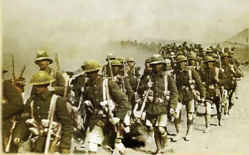 L'image représente un groupe de soldats en marche, probablement pendant une période de conflit. Ils portent des uniformes militaires, y compris des casques et des équipements de combat. Les soldats avancent sur un terrain poussiéreux, et il semble qu'ils soient dans un environnement difficile, avec une atmosphère chargée de fumée ou de poussière. L'image donne une impression de détermination et de camaraderie parmi les troupes.