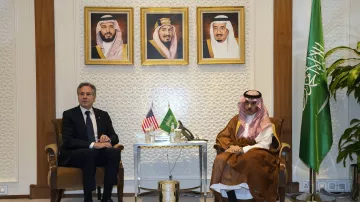 Nell'immagine si possono vedere due uomini seduti uno di fronte all'altro in un ambiente formale. Sullo sfondo ci sono tre ritratti incorniciati, probabilmente di leader sauditi. A sinistra, è presente una bandiera americana e a destra una bandiera saudita. Il tavolo tra i due uomini contiene alcuni oggetti decorativi. L'atmosfera apparente è di dialogo politico o diplomatico.