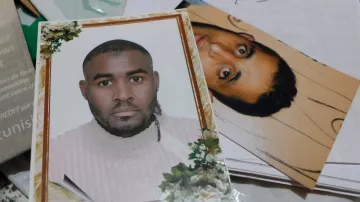 الصورة تحتوي على صورتين شخصيتين. الصورة الأولى لشخص يرتدي سترة بيضاء ويظهر تعبير وجه جدي، محاط بإطار مزخرف بالأزهار. أما الصورة الثانية، فهي لشخص آخر يظهر بشكل غير واضح، مع خلفية بسيطة. يبدو أن الصور موضوعة مع مجموعة من الأوراق أو المستندات.