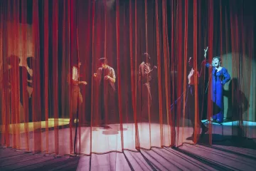 L'image montre une scène de théâtre avec des rideaux en velours rouge et orange qui créent une atmosphère intimiste. On aperçoit plusieurs silhouettes de personnes derrière les rideaux, engagées dans des activités qui semblent dynamiques et expressives. L'un des personnages, visible à droite, porte un costume bleu et semble gesticuler avec enthousiasme. Les jeux de lumière entre les rideaux ajoutent une ambiance dramatique à la scène, accentuant les mouvements des acteurs.