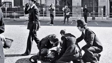L'image montre une scène policière en noir et blanc. Plusieurs policiers en uniforme entourent une personne au sol, visiblement en train d'être arrêté ou assisté. D'autres policiers se tiennent en arrière-plan, certains prenant des notes ou observant la situation. L'environnement semble urbain, avec des barrières de sécurité visibles, probablement autour d'une zone publique. L'atmosphère dégage une impression d'urgence et d'autorité.