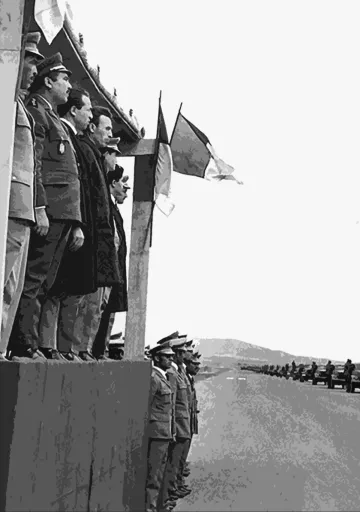 L'image montre un groupe de militaires et de dignitaires, probablement lors d'une cérémonie officielle. Ils se tiennent sur une estrade, certains en uniforme avec des médailles, tandis que d'autres portent des vêtements civils. En arrière-plan, on peut voir des drapeaux, dont un drapeau français, indiquant un contexte lié à la France ou un événement officiel. Les soldats se tiennent en position de parade, face à l'estrade. L'ambiance semble solennelle.