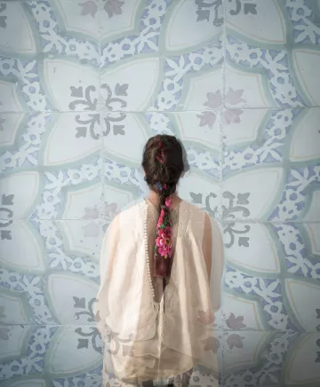 Cette image montre une personne de dos, portant un vêtement léger et délicat. Sa longue tresse, ornée de fleurs colorées, est visible et se détache sur un fond de motifs bleu clair. Les motifs sont ornés de formes géométriques et florales, créant une ambiance apaisante et artistique. L'ensemble évoque une atmosphère de calme et de beauté.