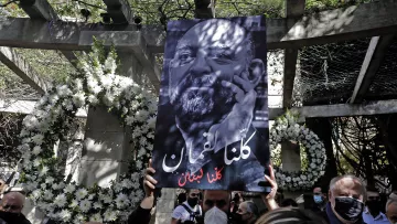 L'immagine mostra una persona che tiene in alto un grande cartello. Il cartello presenta un ritratto in bianco e nero di un uomo con una barba, accompagnato da un testo in arabo che significa "Siamo tutti Libano". Sullo sfondo, ci sono decorazioni floreali e un ambiente che sembra essere un luogo commemorativo. La scena trasmette un senso di unità e solidarietà.