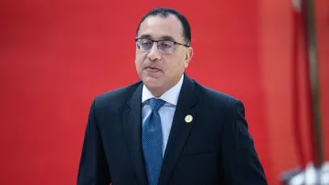 The image features a man dressed in a formal dark suit with a light blue tie. He appears to be in a professional setting, as suggested by the suit and tie. The background is bright red, which adds a striking contrast to his attire. The man looks serious and focused, possibly engaged in an official event or meeting.