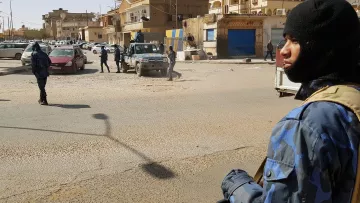 تظهر الصورة مشهداً في منطقة حضرية، حيث يوجد عدد من رجال الأمن يرتدون زيًّا رسميًّا وقبعات سُوداء. أحدهم يحمل سلاحًا ويقف في المقدمة، بينما يتواجد آخرون في الخلفية. تشهد المنطقة حركة مرورية، مع وجود سيارات متوقفة في الشارع. المباني المحيطة تظهر بألوان متنوعة، وبعضها يبدو أنه مغلق. تعكس الصورة أجواءً من اليقظة والحذر الأمني.