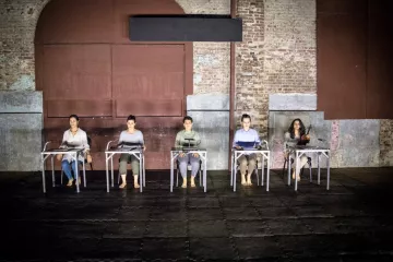 L'image montre un groupe de cinq personnes assises sur des chaises, disposées en ligne. Elles portent des vêtements simples et sont pieds nus, ce qui donne une impression de simplicité et d'intimité. L'environnement est industriel, avec des murs en brique apparente et une lumière tamisée qui crée une ambiance à la fois contemplative et moderne. Les personnes semblent concentrées, chacune avec un objet devant elle, suggérant un moment de performance ou de réflexion partagée.