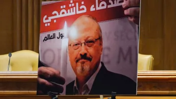 L'image montre un portrait d'un homme tenant une bougie, accompagné d'un texte en arabe et en français. Le fond de l'affiche évoque un hommage ou un soutien, probablement lié à une question de justice. Le contexte global semble être une commémoration ou une manifestation pour défendre les droits humains. Deux chaises sont visibles en arrière-plan, suggérant que l'affiche est présentée lors d'un événement formel.