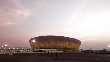 L'image montre un grand stade moderne à l'architecture distinctive, situé sous un ciel teinté de nuances pastel au coucher du soleil. Le stade possède une structure en forme d'ellipse avec une façade brillante, peut-être en métal ou en verre, qui capte la lumière ambiante. On aperçoit quelques personnes marchant devant le stade, soulignant sa taille imposante. L'ensemble de la scène dégage une ambiance calme et majestueuse.
