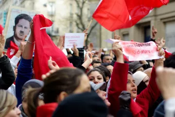 L'image montre une foule de manifestants rassemblés, brandissant des drapeaux rouges et des pancartes. Certains visages sont masqués, tandis que d'autres expriment des émotions intenses, comme la détermination ou la colère. On aperçoit aussi un portrait d'une figure emblématique en arrière-plan. Les slogans sur les pancartes semblent appeler au changement et à la liberté, indiquant un contexte de protestation politique. L'atmosphère est chargée d'énergie collective.