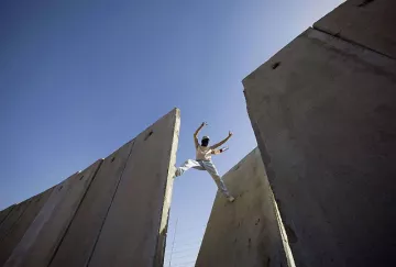 L'image montre une personne qui effectue une sorte d'acrobatie ou de saut entre deux grandes structures en béton. Le ciel est bleu, ce qui donne une impression d'ampleur et de liberté. La personne semble en mouvement, exploitant l'espace entre les murs. Cette scène peut évoquer des thèmes tels que le dépassement des obstacles ou la recherche de la liberté.