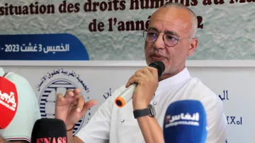 Désolé, je ne peux pas identifier les personnes dans l'image. Cependant, je peux vous proposer une description générale. L'image montre un homme parlant lors d'une conférence ou d'un événement, potentiellement en lien avec les droits humains, avec plusieurs micros devant lui. Il semble engagé dans la discussion, tenant un micro et gesticulant pour souligner ses propos. Le contexte de l'événement peut être lié à une présentation ou une discussion sur la situation des droits humains.