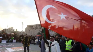 L'image montre une scène de rassemblement ou de manifestation en plein air. Au premier plan, un homme âgé tient fièrement un grand drapeau turc, qui est rouge avec une étoile et un croissant blancs. On peut apercevoir une foule de personnes en arrière-plan, certaines portant des vêtements de sécurité fluorescents. La scène se déroule peut-être dans un lieu historique, avec un bâtiment ancien visible derrière la foule. L'ambiance semble dynamique et engagée.