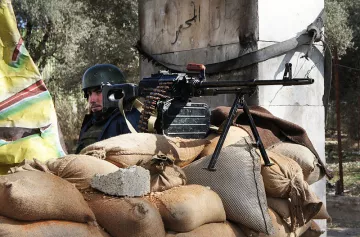 L'image montre un soldat en arrière-plan, vêtu d'un uniforme et portant un casque. Devant lui, il y a une mitrailleuse montée sur un trépied, posée sur un tas de sacs de sable. Le contexte semble être une zone de conflit, avec des éléments de construction en arrière-plan, probablement dans une région touchée par des combats. La scène évoque des mesures de protection et de défense.