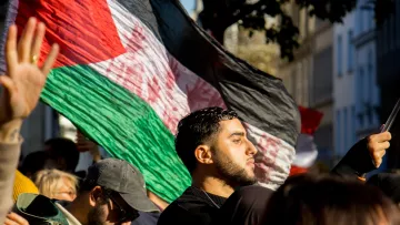 در این تصویر، گروهی از افراد در حال برگزاری یک تظاهر یا تجمع هستند. یک مرد جوان در جلو تصویر دیده می‌شود که به پرچم کشور خود (که احتمالاً پرچم فلسطین است) افتخار می‌کند. او با جدیت به جلو نگاه می‌کند. در پس‌زمینه، تعدادی از افراد دیگر نیز حضور دارند که برخی از آن‌ها کلاه به سر دارند و به نظر می‌رسد که در حال حمایت از یک هدف مشترک هستند. نور آفتاب به تصویر زیبایی لطافتی می‌بخشد و فضای تجمع را گرم و زنده نشان می‌دهد.