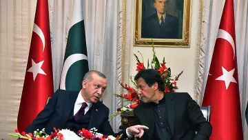 En la imagen se pueden ver a dos hombres conversando en un ambiente formal, rodeados de banderas de Turquía y Pakistán. Hay un arreglo floral colorido en la mesa, y detrás de ellos se encuentra un retrato en la pared. La escena transmite un ambiente de diplomacia y colaboración entre los dos países.