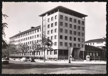 L'image représente un bâtiment architectural de plusieurs étages, probablement de style moderne. Il est photographié sous une lumière naturelle, mettant en avant sa façade aux lignes géométriques. Le bâtiment semble être entouré d'arbres et de quelques véhicules anciens, ce qui donne une ambiance d'époque. On peut également apercevoir des éléments de la rue, comme des lampadaires et éventuellement d'autres bâtiments en arrière-plan. L'ensemble de l'image présente une esthétique monochrome, accentuant le caractère historique de la scène.