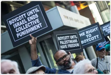 The image depicts a group of protesters holding signs at a demonstration. Some of the signs read "BOYCOTT UNTIL ISRAEL ENDS COLLECTIVE PUNISHMENT" and "BOYCOTT UNTIL PALESTINIANS HAVE FREEDOM." The atmosphere suggests a call for social justice and awareness regarding the Palestinian situation. The protesters appear engaged and determined in their stance.