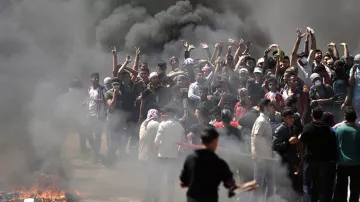 L'image montre un groupe de personnes rassemblées dans une atmosphère tendue. Elles sont entourées de fumée noire, probablement causée par des feux. Certains manifestants lèvent les bras en signe de protestation. On peut sentir une ambiance de colère et de résistance, avec quelques personnes qui semblent brandir des objets. Le décor est chaotique, évoquant une situation de crise ou de manifestation.