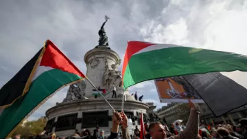 تظهر الصورة مجموعة من الأشخاص وهم يحملون أعلام فلسطينية في تجمع جماهيري. الخلفية تُظهر نصبًا تذكاريًا عظيمًا، مع سماء ملبدة بالغيوم. يبدو أن الناس متحمسون ويشاركون في فعالية ذات طابع وطني أو سياسي. الأعلام تحمل ألوان العلم الفلسطيني، مما يشير إلى دعمهم للقضية الفلسطينية.
