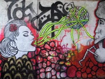 L'image montre une œuvre de street art colorée sur un mur. À gauche, il y a le portrait d'une femme avec un rose dans les cheveux, qui semble "parler" ou "chanter". À droite, un homme est représenté, affichant une expression sérieuse. Entre les deux, des lignes vertes émanent de la tête de la femme, semblant évoquer une connexion ou un échange de pensées, illustrées par un cerveau stylisé en arrière-plan. L'ensemble est dominé par des couleurs vives et des motifs abstraits, créant une atmosphère vibrante et dynamique.