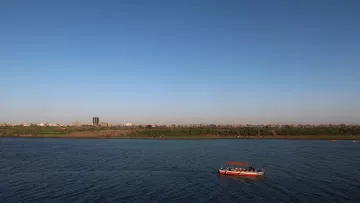 تظهر في الصورة منظرًا طبيعيًا جميلًا يتضمن نهرًا هادئًا ينساب تحت سماء صافية وزرقاء. على جانب النهر، يمكن رؤية مجموعة من الأشجار والنباتات التي تزين الساحل. بينما في وسط النهر، تبحر قارب صغير ذو سقف برتقالي، حيث يبدو أن هناك أشخاصًا يستمتعون بالرحلة. في الخلفية، تمتد المدينة مع المباني السكنية، مما يضفي طابعًا حضريًا على المشهد الطبيعي.