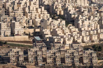 L'image présente un paysage urbain dense, constitué de nombreux bâtiments aux formes géométriques. Les structures sont principalement de couleur claire, avec des nuances de beige et de blanc. On peut observer des immeubles empilés les uns sur les autres, créant une apparence presque uniforme. Le terrain autour des bâtiments semble légèrement vallonné, avec des zones de végétation sporadique. Il y a également des éléments architecturaux distincts, tels qu'un toit qui se distingue parmi l'ensemble des constructions. L'ensemble donne une impression de modernité et d'urbanisation.