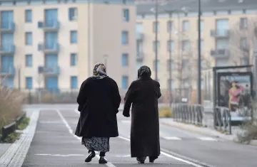 L'image montre deux femmes marchant côte à côte sur une voie urbaine. Elles portent des manteaux sombres et des foulards, ce qui suggère qu'il fait frais. En arrière-plan, on distingue des bâtiments modernes avec des balcons et des fenêtres. La scène dégage une atmosphère tranquille, avec un chemin bordé d'herbes. Leurs silhouettes semblent illustrer un moment de complicité et de camaraderie.