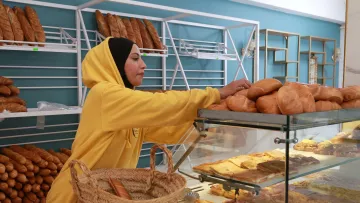 L'image montre une femme dans une boulangerie. Elle porte un sweat à capuche jaune et est en train de disposer des pains sur une étagère vitrée. La boulangerie est bien équipée avec une variété de pains et de pâtisseries visibles derrière le verre. Il y a également un panier en osier sur le comptoir. L'ambiance est chaleureuse et invitante, avec des couleurs douces et un éclairage agréable.