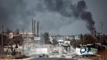 The image depicts a desolate street under a dark, smoke-filled sky. In the background, billowing plumes of black smoke suggest an ongoing fire or explosion. The foreground shows a damaged road with debris, potentially hinting at a conflict zone. There are signs of destruction, including burnt vehicles and abandoned structures, contributing to an overall atmosphere of turmoil and devastation. The landscape appears barren with a few power lines and distant buildings, reflecting the impact of violence on the area.
