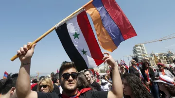 L'image montre un groupe de personnes participant à une manifestation ou un rassemblement. Un homme au premier plan tient fièrement trois drapeaux : le drapeau syrien, un drapeau avec trois étoiles vertes sur fond noir et rouge, ainsi qu'un drapeau arménien. L'ambiance semble festive et engagée, avec de nombreuses personnes en arrière-plan, probablement partageant des sentiments communs ou soutenant une cause. Le ciel est clair, ce qui ajoute à l'énergie de l'événement.