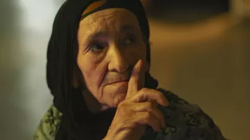 L'image montre une femme âgée avec un regard pensif. Elle porte un foulard noir qui encadre son visage. Son expression est sérieuse et elle semble faire un geste de silence avec son doigt posé sur ses lèvres. La lumière dans le fond est douce, créant une ambiance calme et introspective. Les détails de son visage reflètent une certaine sagesse et vécu.