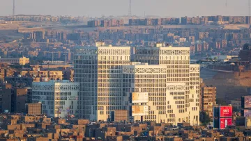 L'image montre un paysage urbain avec plusieurs bâtiments modernes au premier plan, qui se distinguent par leur architecture unique et leurs façades vitrées. En arrière-plan, on aperçoit une vaste étendue de constructions résidentielles plus anciennes, avec des habitations compactes et souvent uniformes. La lumière du soleil semble éclairer les bâtiments modernes, créant un contraste avec le reste de la ville, particulièrement à l'heure dorée du matin ou du soir. Le ciel apparaît clair, ce qui met en valeur la scène urbaine.