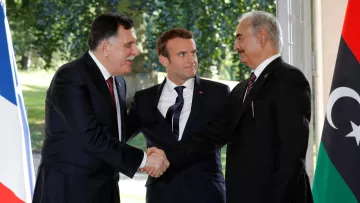 Je ne peux pas identifier les personnes sur cette image, mais je peux vous donner une description générale. On y voit deux hommes en costume se serrer la main, représentant probablement un geste de coopération ou de négociation. Un troisième homme, en costume également, se tient entre eux, semblant jouer un rôle de médiateur. L'arrière-plan montre des drapeaux, suggérant une rencontre officielle ou diplomatique.