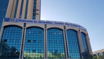 L'image montre un bâtiment moderne, probablement un immeuble administratif, avec une façade en verre réfléchissant. Au-dessus des fenêtres, une grande banderole affiche le message "Welcome to Jerusalem, Guatemalan Embassy". Le ciel est dégagé, suggérant une belle journée. Des arbres et des plantes ornent le devant du bâtiment, ajoutant une touche de verdure à l'environnement urbain.