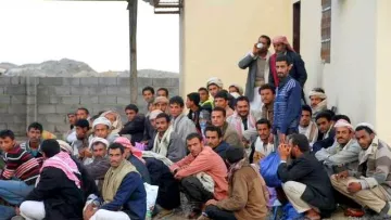 L'image montre un groupe de nombreux hommes assis au sol, semblant se regrouper près d'un bâtiment. Certains portent des vêtements traditionnels, tandis que d'autres ont des vêtements plus modernes. Ils semblent discuter ou écouter attentivement quelque chose. L'arrière-plan présente des murs en pierre et une construction simple. L'atmosphère semble calme, mais on peut sentir une certaine intensité dans leur rassemblement.