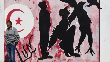 L'image montre un mur peint avec des graffitis. À gauche, on peut voir le drapeau tunisien en rouge et blanc, avec un croissant et une étoile. À côté, des silhouettes humaines sont représentées en noir sur un fond rose, certaines semblent interagir avec des éléments d'oiseaux, suggérant des thèmes de liberté ou de mouvement. Un homme en pull rayé marche le long du mur, ajoutant un aspect vivant à la scène.