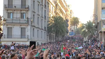 این تصویر نمایی از یک اجتماعات بزرگ در خیابانی شلوغ را نشان می‌دهد. مردم زیادی در حال تظاهرات یا جمع شدن هستند و در میان آن‌ها تعدادی پرچم به چشم می‌خورد. درختان بلند نخل در دو طرف خیابان دیده می‌شوند و ساختمان‌های مرتفع با بالکن‌ها نیز در پس‌زمینه قرار دارند. مردم با entusiasmo در حال برنداشتن تلفن‌های همراه خود و ثبت این لحظه هستند. جو عمومی تصویری از شور و شوق و همبستگی را القا می‌کند.