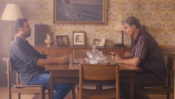 L'image montre deux hommes assis à une table en bois dans une pièce décorée avec un papier peint aux motifs rétro. L'ambiance est intérieure et intimiste, avec une lampe à côté et des photos accrochées au mur. Les deux hommes semblent engagés dans une conversation sérieuse. Le premier homme, à gauche, est vêtu d'une chemise sombre et regarde l'autre. Le deuxième homme, à droite, fume une cigarette et semble pensif. L'éclairage doux ajoute à l'atmosphère.