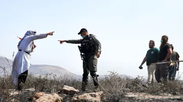 L'image montre une scène de tension entre un homme habillé dans un style traditionnel, tenant une sorte de bâton ou d'objet, qui semble pointer du doigt. Un policier ou un soldat, portant un uniforme et un gilet, est en position d'affrontement avec lui, pointant également vers l'homme. En arrière-plan, plusieurs autres personnes, visiblement des observateurs ou des participants à la scène, se tiennent. Le paysage environnant semble aride, avec des rochers. L'atmosphère est chargée et indique une situation potentiellement conflictuelle.