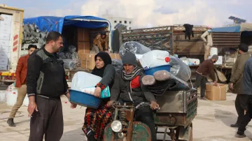 L'image montre une scène de marché ou de distribution dans un environnement rural. On y voit un homme et une femme discutant, tandis qu'un autre homme est assis sur un véhicule à trois roues, probablement en train de transporter des marchandises. Le décor entoure des containers en plastique, et plusieurs personnes s'affairent à décharger ou à déplacer des articles. Le ciel est nuageux, suggérant une atmosphère d'activité et d'échange.